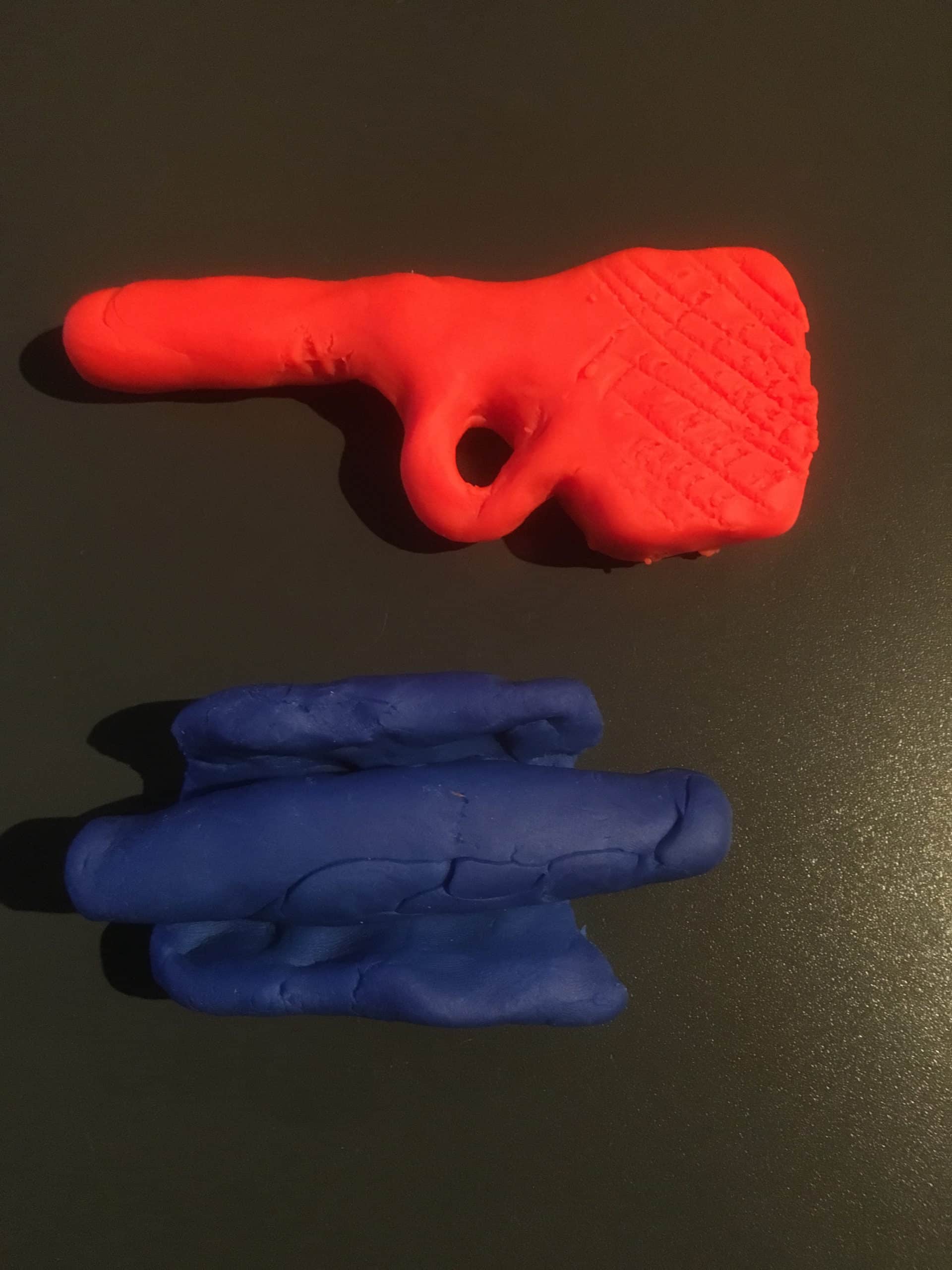 Två figurer i färgad lera ligger på en mörk yta. En röd figur som kan tolkas som en pinne med en ögla på samt en blå figur i form av ett rör med flikar uppe och nere.