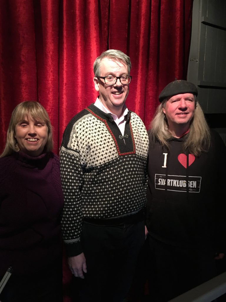 Anna, Richard och Ulf, gruppfoto med röda gardiner som bakgrund. Anna har långt blont hår och en lila polotröja. Richard har kort ljust hår, glasögon och en svart och beige tröja med prickigt mönster och metallspänne över kragen framtill. Ulf har en svart basker, långt grått hår och en svart munktröja med texten: I love Svartklubben.