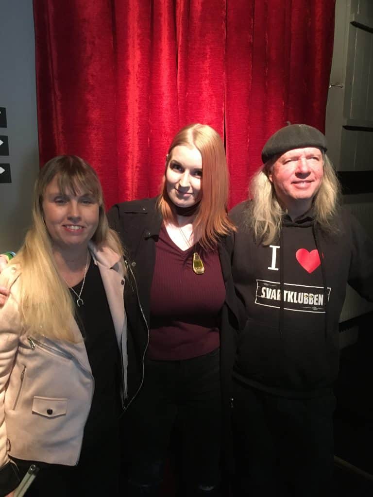 Anna, Johanna och Ulf, gruppfoto med röda gardiner som bakgrund. Anna har långt blont hår, ljus kavajjacka och svart tröja under. Johanna har långt rött hår, lila undertröja och en svart tröja eller jacka ovanpå. Ulf har grått långt hår, svart basker, svart huvtröja med texten: I love Svartklubben.