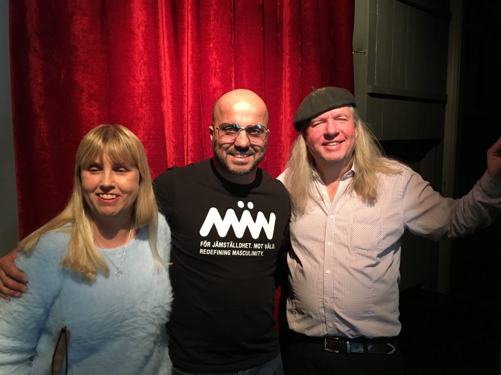 Anna, Alán och Ulf, gruppfoto med röda gardiner som bakgrund. Anna har långt blont hår och en ljusblå tjock tröja. Alán har rakat huvud, glasögon och en svart tröja med texten: Män, För jämställdhet, mot våld. Redefining masculinity