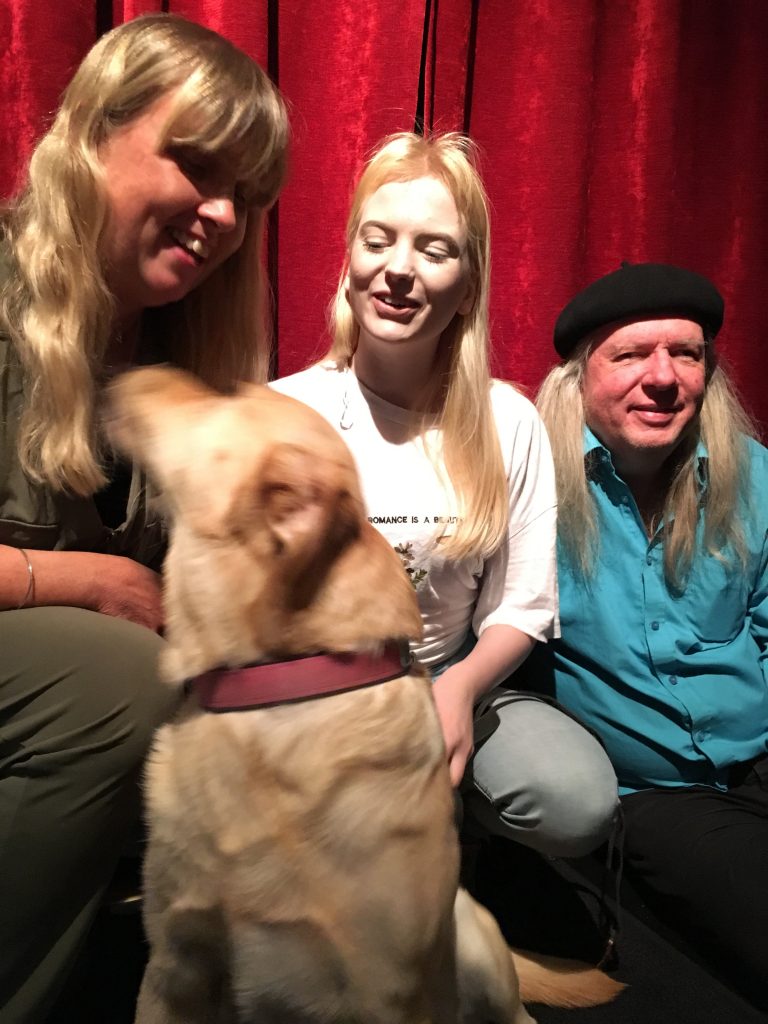 Anna, Smilla och Ulf med hunden Chanti, en ljus labrador. Anna har långt blont hår och en grön byxdress. Smilla har långt blont hår, vit t-shirt och ljusblå jeans. Ulf har en svart basker, långt grått hår, turkos skjorta och mörka byxor.