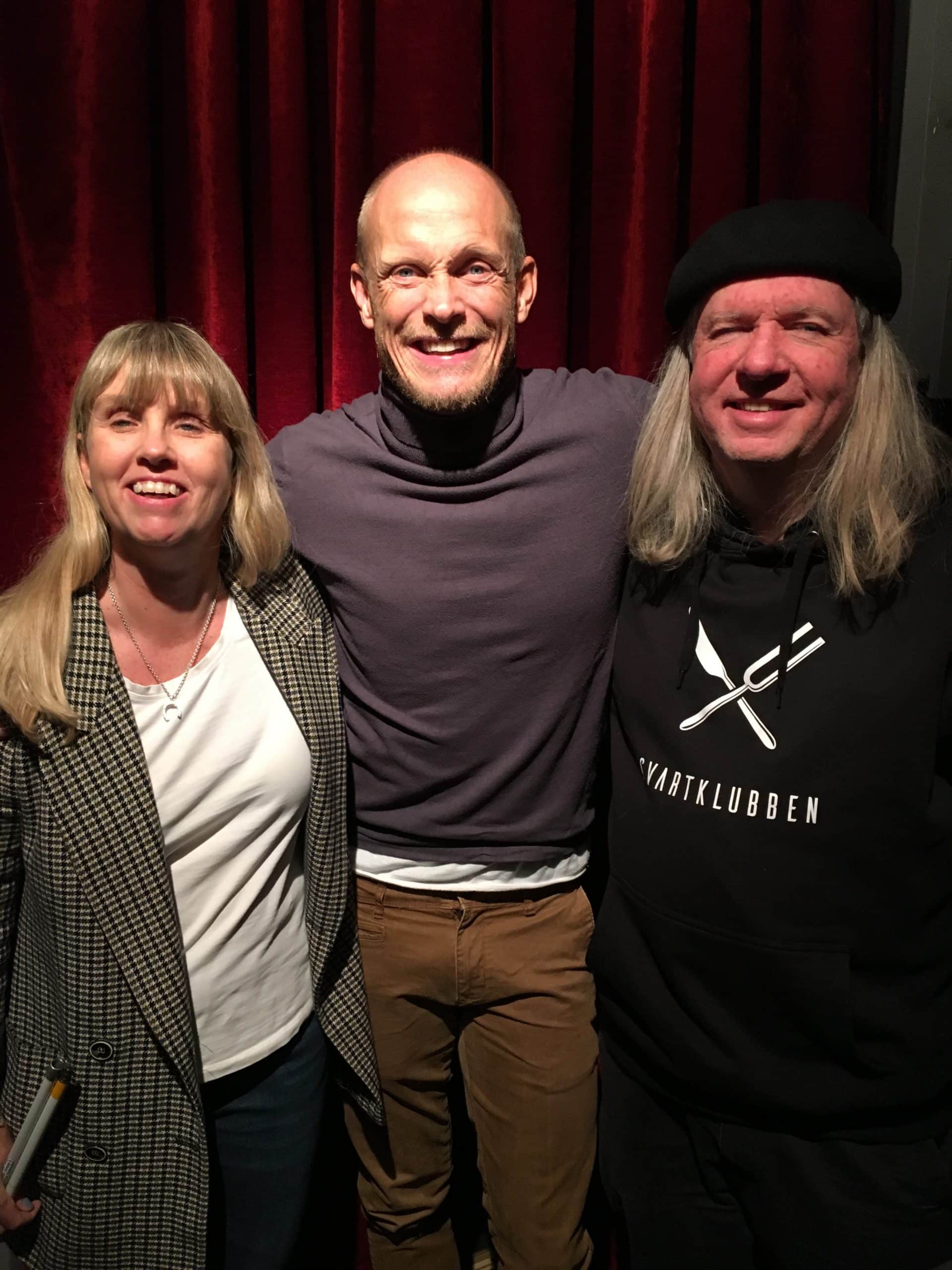 Anna, Tobias och Ulf, gruppfoto med röda gardiner som bakgrund. Anna har långt blont hår och har på sig en gråmönstrad jacka, vit tröja och mörka byxor. Tobias har rakat huvud, skägg, har på sig en lila polotröja, bruna jeans. Ulf har grått långt hår, svart basker, svart huvtröja med Svartklubbens logotyp