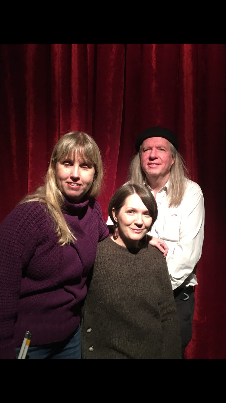 Anna, Yasmin och Ulf gruppfoto med röda gardiner som bakgrund. Anna har långt blont hår och har på sig en lila långärmad tröja. Yasmin sitter ned och har på sig en långärmad gråbrun tröja samt halvlångt brunt hår. Ulf har långt grått och, vit skjorta och mörka byxor.