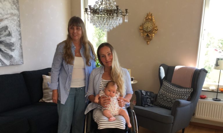 Anna och Charlotte hemma i Charlotte's vardagsrum. Anna står bakom Charlotte med långt blont hår och lugg framtill samt iklädd blårandig långärmad skjorta, vit topp och ljusblå jeans och står barfota. Charlotte sitter i sin rullstol. Hon har långt blont hår, ljusblå långärmad skjorta, vit kjol med svarta streck samt svarta leggings och vita skor. I knäet har hon sin nyfödda Alicia. I bakgrunden syns en mörk soffa, grå fåtölj, kristallkrona och en guldklocka på väggen.