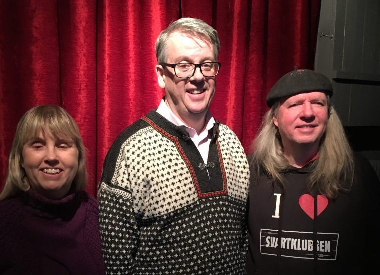 Anna, Richard och Ulf, gruppfoto med röda gardiner som bakgrund. Anna har långt blont hår och en lila polotröja. Richard har kort ljust hår, glasögon och en svart och beige tröja med prickigt mönster och metallspänne över kragen framtill. Ulf har en svart basker, långt grått hår och en svart munktröja med texten: I love Svartklubben.