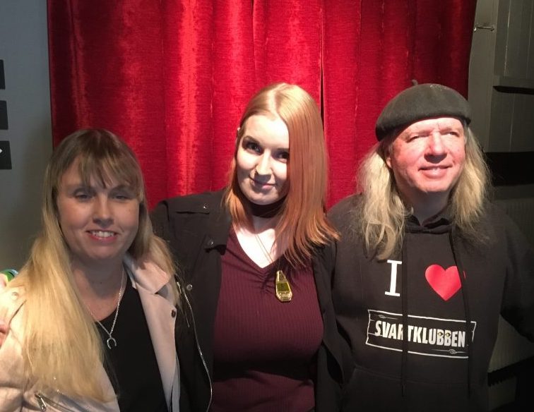 Anna, Johanna och Ulf, gruppfoto med röda gardiner som bakgrund. Anna har långt blont hår, ljus kavajjacka och svart tröja under. Johanna har långt rött hår, lila undertröja och en svart tröja eller jacka ovanpå. Ulf har grått långt hår, svart basker, svart huvtröja med texten: I love Svartklubben.