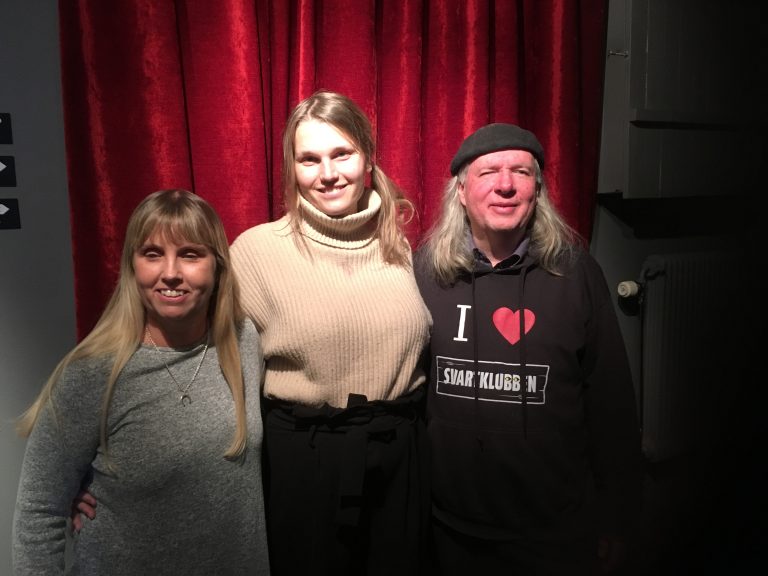 Anna, Aleksa och Ulf, gruppfoto med röda gardiner som bakgrund. Anna har långt blont hår och har på sig en grå långärmad tröja. Aleksa har långt mörkblont hår och på sig en beige tjock polotröja samt mörk klädsel på benen. Ulf har på sig en svart basker, långt grått hår samt en svart munkjacka med texten: I love Svartklubben.