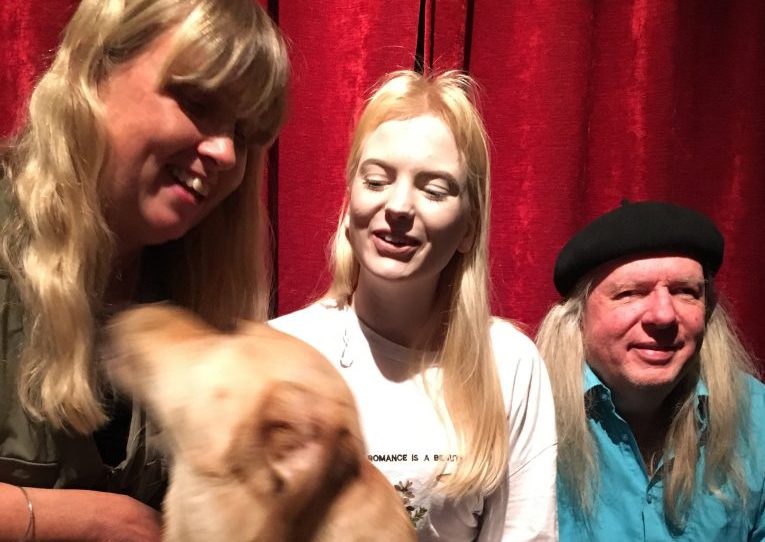 Anna, Smilla och Ulf med hunden Chanti, en ljus labrador. Anna har långt blont hår och en grön byxdress. Smilla har långt blont hår, vit t-shirt och ljusblå jeans. Ulf har en svart basker, långt grått hår, turkos skjorta och mörka byxor.