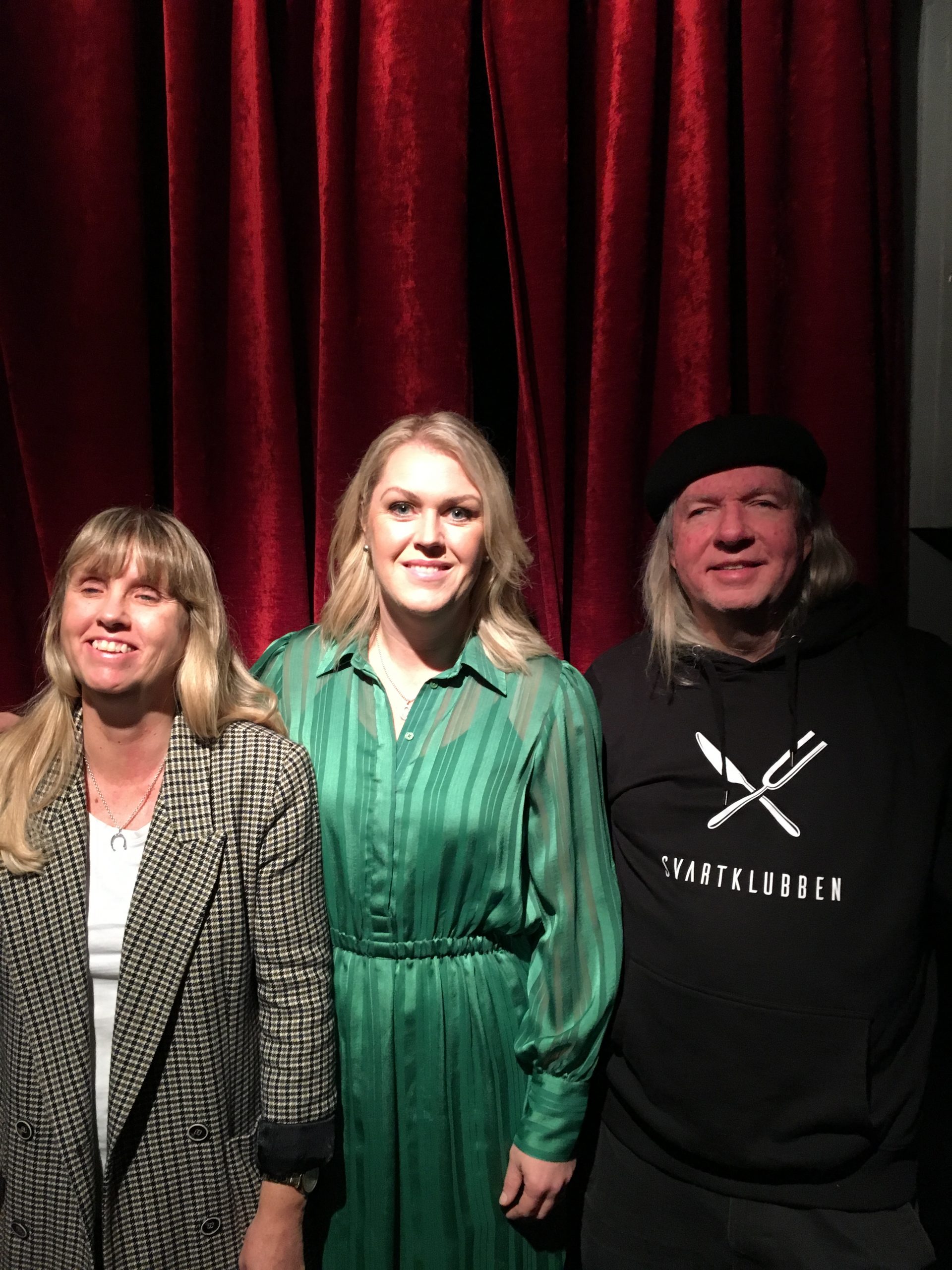 Anna, Lena och Ulf, gruppfoto med röda gardiner som bakgrund. Anna har långt blont hår och har på sig en gråmönstrad jacka och vit undertröja. Lena har på sig en ljusgrön randig klänning och långt blont hår. Ulf har grått långt hår, svart basker, svart huvtröja med Svartklubbens logotyp.