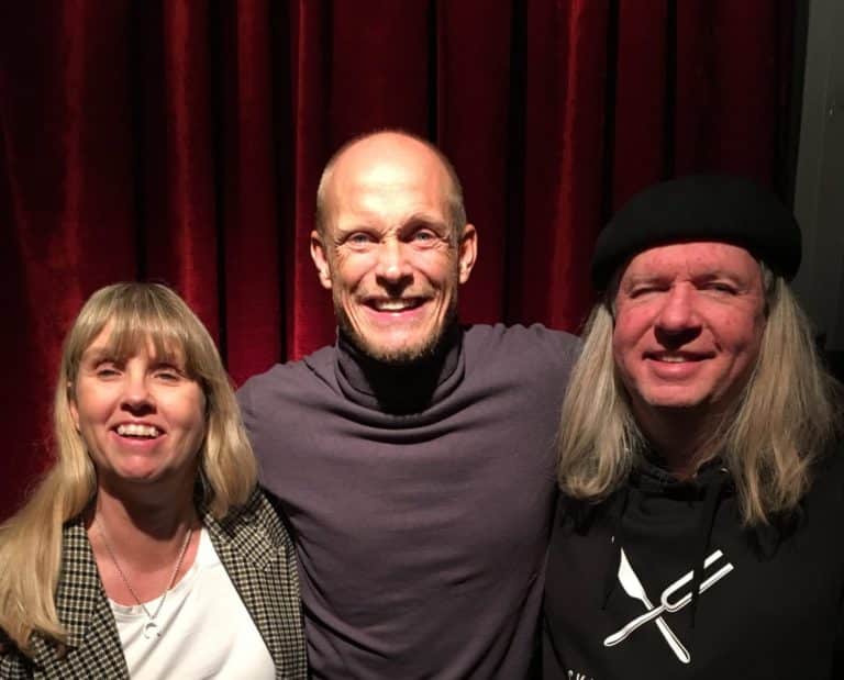 Anna, Tobias och Ulf, gruppfoto med röda gardiner som bakgrund. Anna har långt blont hår och har på sig en gråmönstrad jacka, vit tröja och mörka byxor. Tobias har rakat huvud, skägg, har på sig en lila polotröja, bruna jeans. Ulf har grått långt hår, svart basker, svart huvtröja med Svartklubbens logotyp