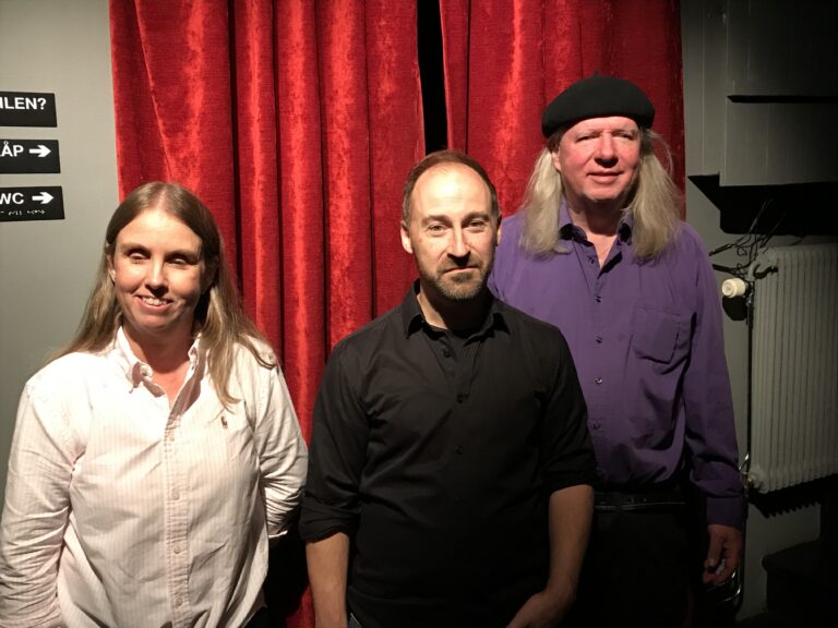 Anna, Johan och Ulf, gruppfoto i studion. Anna till vänster står med vit skjorta, långt blont hår. Johan har kort brunt hår och stubbigt skägg samt svart skjorta med uppkavlade ärmar. Ulf till höger i svart basker, långt grått hår, långärmad lila skjorta. I bakgrunden ett rött drapperi mot gråvit vägg.