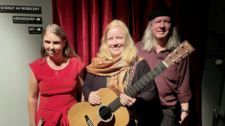 Anna, Tina och Ulf står i studion. Anna har blont långt utsläppt hår, röd kjol/klänning, Tina ljusblont långt hår, gulmönstrad scarf, mörk långärma tröja och håller i en gulbrun gitarr. Ulf tlll höger i svart basker, långt grått hår, vinröd skjorta, mörka byxor. I bakgrunden ett rött drapperi.