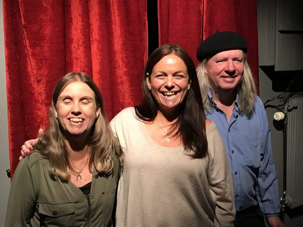 Anna, Katarina och Ulf, gruppfoto i studion. Anna till vänster står med grön långärmad skjorta, långt blont hår. Katarina har långt brunt hår och en vit långärmad tröja. Ulf till höger i svart basker, långt grått hår, långärmad ljusblå skjorta. I bakgrunden ett rött drapperi mot gråvit vägg.