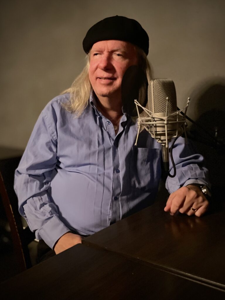 Ulf sitter bakom en mikrofon i studion och ler. Han har på sig en basker och ljusblå skjorta
